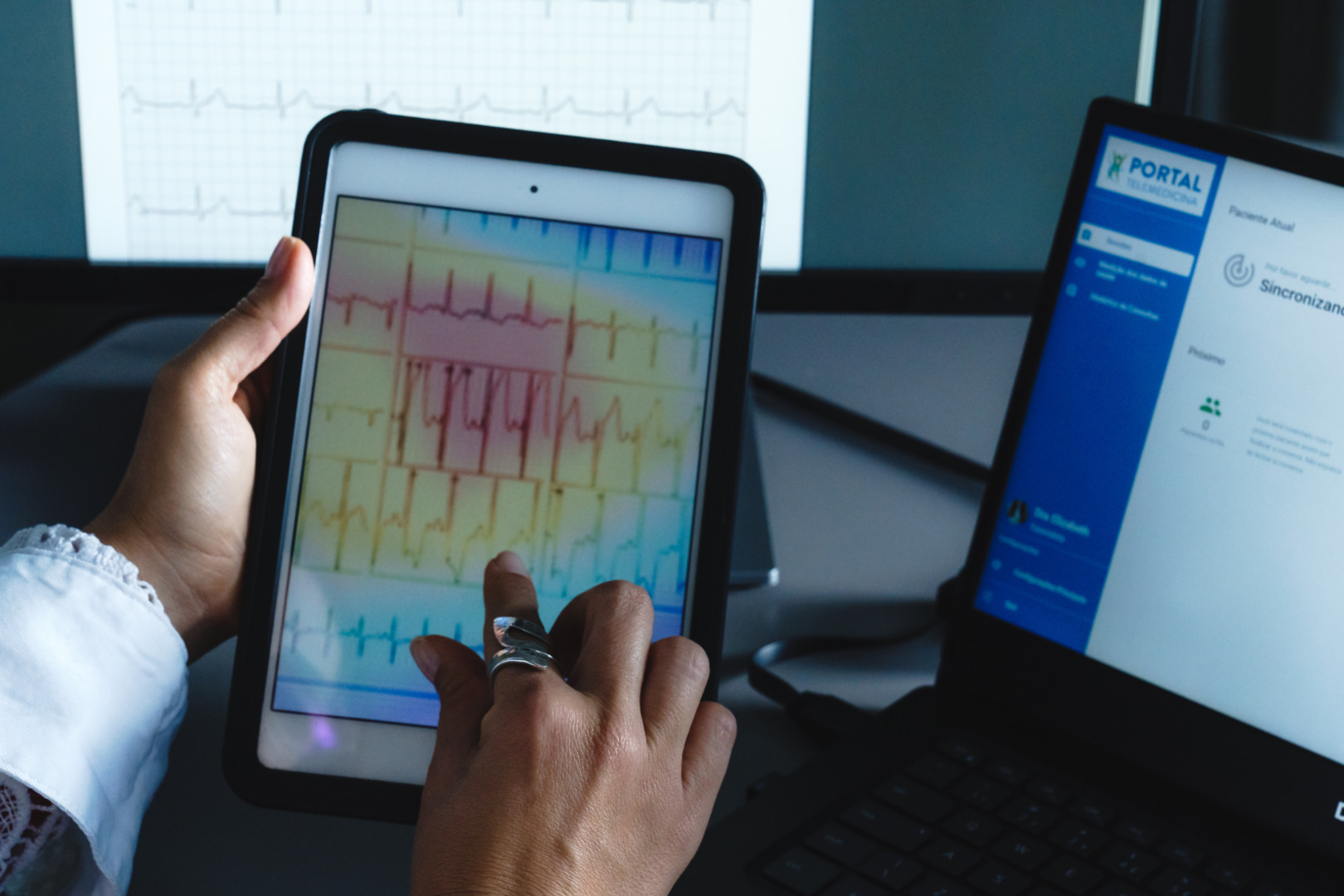 Healthcare professional analyzing an ECG on a tablet with a laptop displaying medical data in the background