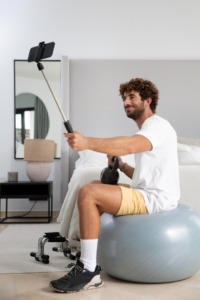 homem jovem fazendo exercício em bola de pilates e utilizando celular em suporte 