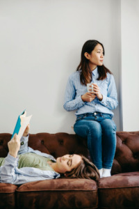 mulher sentada no encosto do sofá e outra mulher deitada lendo livro