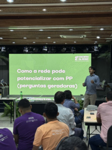 homem em pé apresentando slides em telão para pessoas assistindo