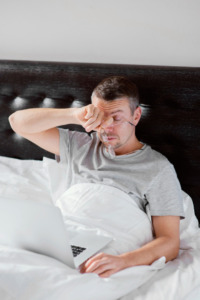 homem sentado na cama, demonstrando cansaço, com notebook no colo