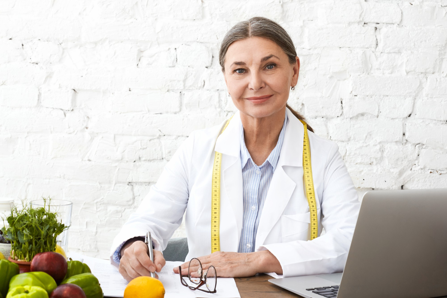 nutricionista em consultório, com fita métrica de medidas no pescoço e alimentos na mesa