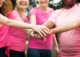 mulheres em pé, em área aberta, com camisetas rosa e colocando uma mão sobre a outra