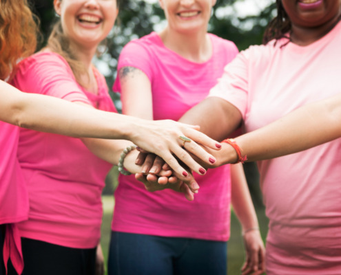 mulheres em pé, em área aberta, com camisetas rosa e colocando uma mão sobre a outra