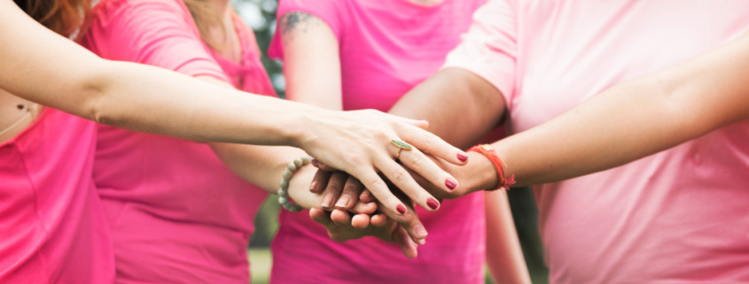 mulheres em pé, em área aberta, com camisetas rosa e colocando uma mão sobre a outra