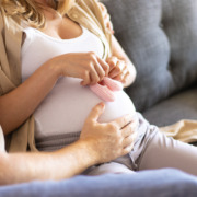 mulher grávida sentada no sofá, ao lado de parceiro que apoia a mão em sua barriga