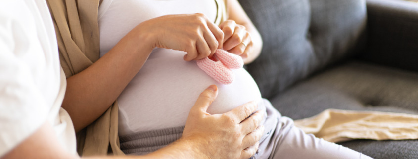 mulher grávida sentada no sofá, ao lado de parceiro que apoia a mão em sua barriga