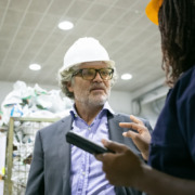 homem de blazer e camisa social, com capacete de segurança em área de produção conversando com funcionária
