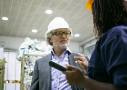 homem de blazer e camisa social, com capacete de segurança em área de produção conversando com funcionária