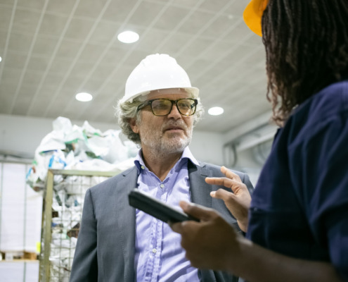 homem de blazer e camisa social, com capacete de segurança em área de produção conversando com funcionária