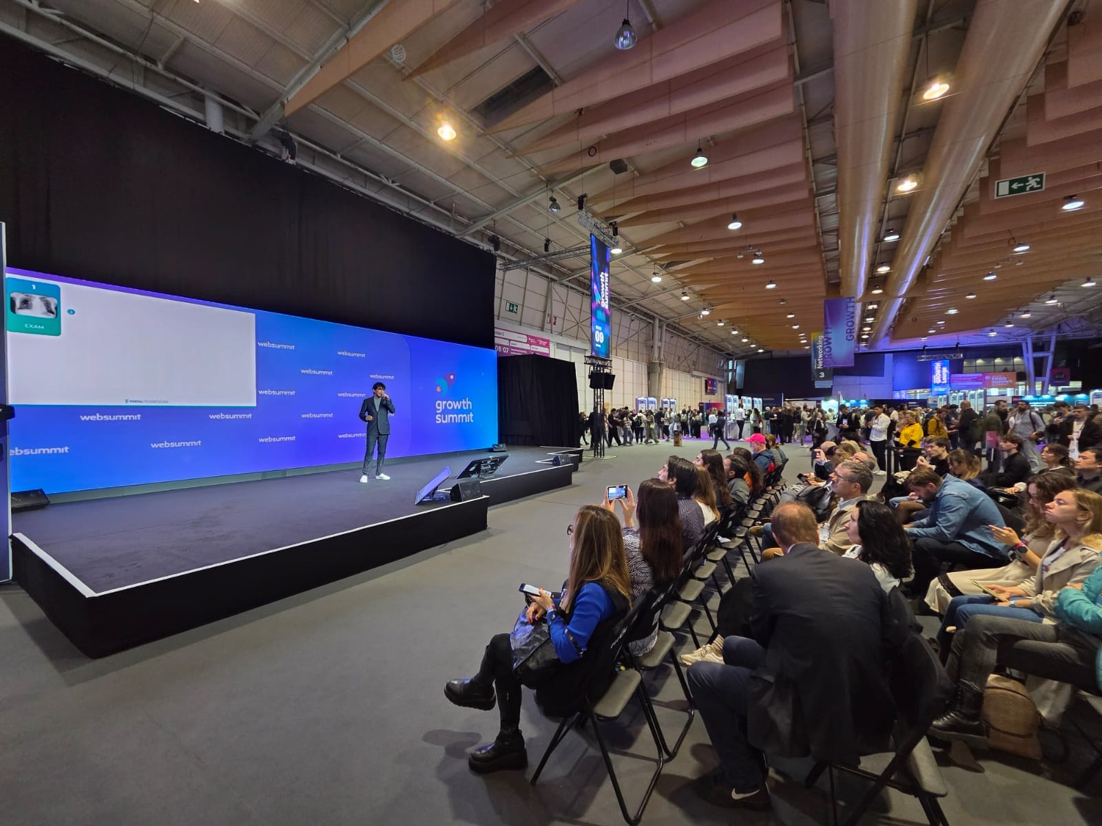 palestrante no palco e público assistindo ao vivo em cadeiras
