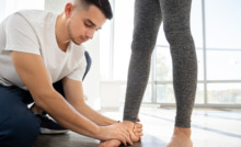 Homem de camiseta branca agachado no chão, avaliando o pé direito de uma mulher em pé