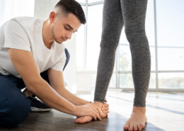 Homem de camiseta branca agachado no chão, avaliando o pé direito de uma mulher em pé