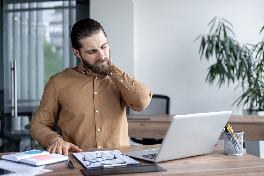 homem sentado em mesa de escritório com mão no pescoço, indicando dor