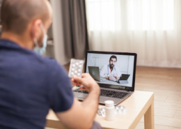 homem em casa segurando cartela de comprimido em consulta no notebook