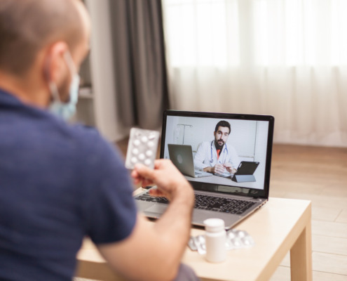 homem em casa segurando cartela de comprimido em consulta no notebook