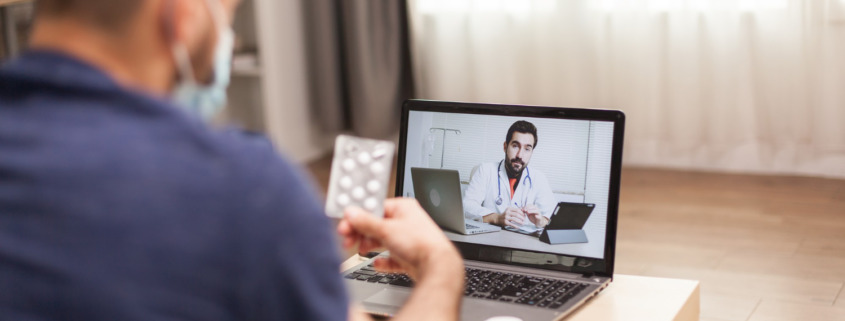 homem em casa segurando cartela de comprimido em consulta no notebook
