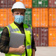 funcionário usando colete luminoso, capacete e máscara em área de porto com containers ao fundo