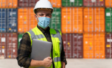 funcionário usando colete luminoso, capacete e máscara em área de porto com containers ao fundo