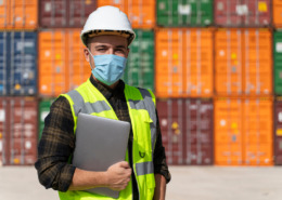 funcionário usando colete luminoso, capacete e máscara em área de porto com containers ao fundo