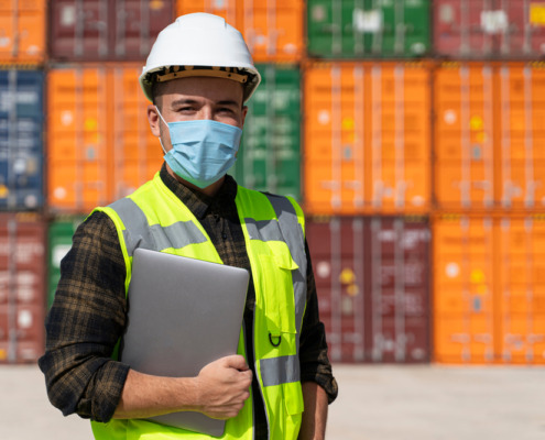 funcionário usando colete luminoso, capacete e máscara em área de porto com containers ao fundo