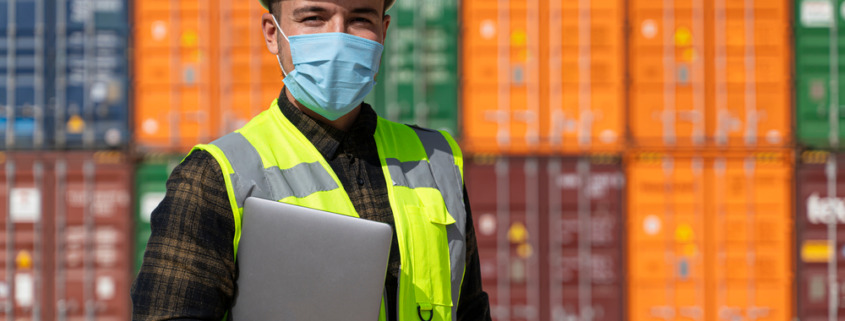 funcionário usando colete luminoso, capacete e máscara em área de porto com containers ao fundo