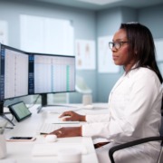 médica em mesa de trabalho analisando dados em dois monitores em sua frente