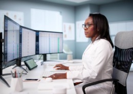 médica em mesa de trabalho analisando dados em dois monitores em sua frente