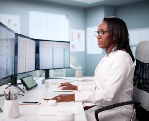 médica em mesa de trabalho analisando dados em dois monitores em sua frente