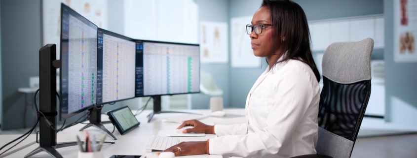 médica em mesa de trabalho analisando dados em dois monitores em sua frente