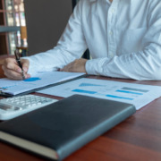 braços de homem de camisa social branca em mesa de trabalho com papeis e agenda na mesa, fazendo contabilidade