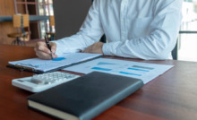 braços de homem de camisa social branca em mesa de trabalho com papeis e agenda na mesa, fazendo contabilidade