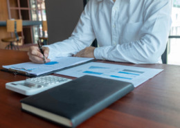 braços de homem de camisa social branca em mesa de trabalho com papeis e agenda na mesa, fazendo contabilidade