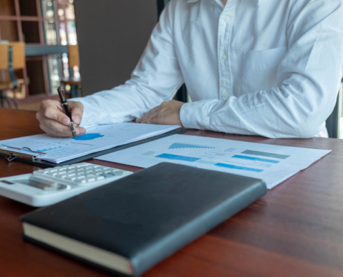 braços de homem de camisa social branca em mesa de trabalho com papeis e agenda na mesa, fazendo contabilidade
