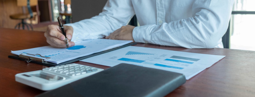 braços de homem de camisa social branca em mesa de trabalho com papeis e agenda na mesa, fazendo contabilidade