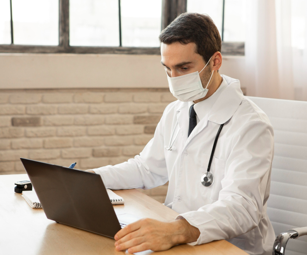 médico em mesa de consultório utilizando máscara facial e notebook