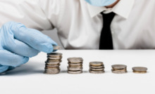 Pessoa com máscara e proteção e luva descartável colocando moedas na mesa indicando economia